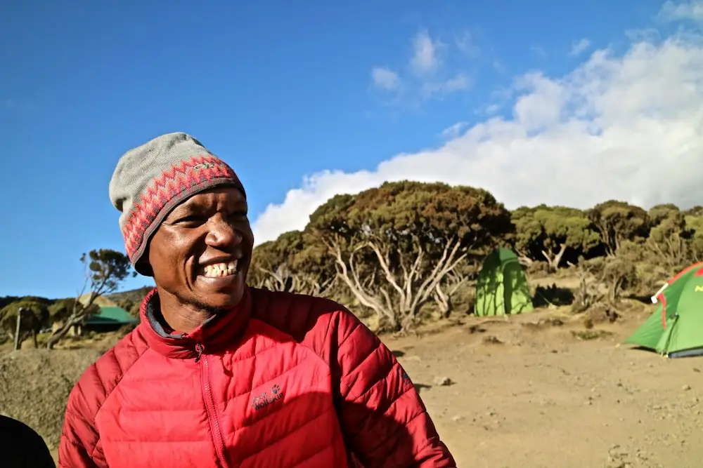 A smiling man in a red jacket and wool hat at a mountain camp